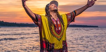 Man in colorful traditional attire with arms raised at sunset beach.