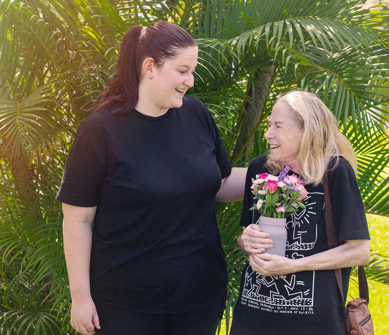 Two women smiling, one holding a bouquet of flowers, standing outdoors with green foliage.