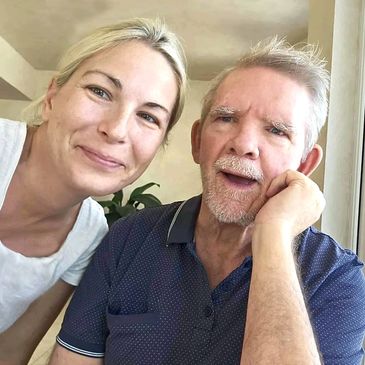 Smiling woman taking a selfie with an elderly man sitting by a window.