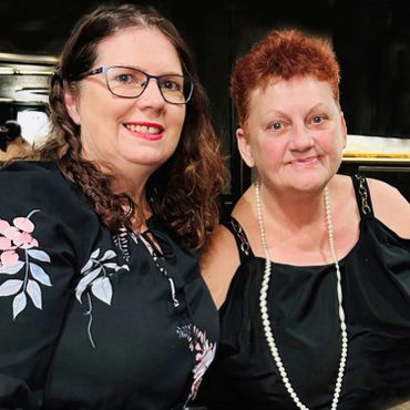 Two women smiling together, one wearing glasses and a floral top, the other with short red hair and a pearl necklace.