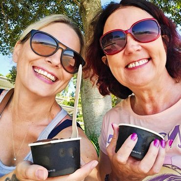 Two women smiling with sunglasses, holding black dessert cups outdoors.