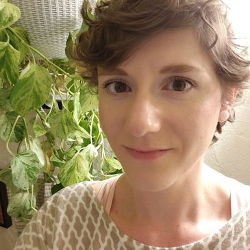 A woman with short curly hair smiling in front of a leafy plant indoors.