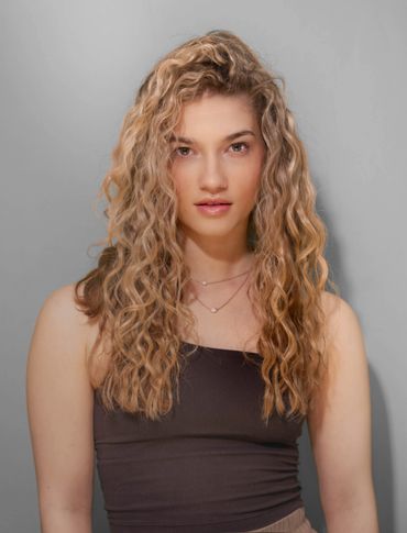 Young woman with curly blonde hair and a black top posing against a gray background.