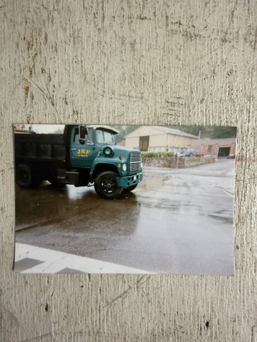 Green JRF dump truck parked on wet pavement near industrial buildings.