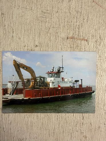 A workboat carrying a yellow excavator on the water.
