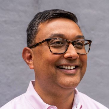 Smiling man wearing tortoiseshell glasses and a pink shirt.