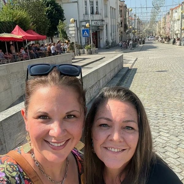 Two women smiling for a selfie on a sunny street with historic buildings and people in the background.