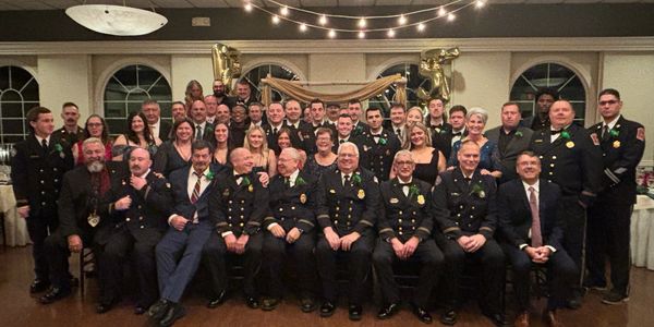 Group photo of firefighters and guests at an indoor event decorated with string lights and balloons.