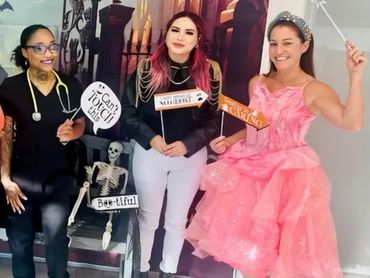 Group of women in Halloween costumes holding festive signs and a dog, posing indoors.