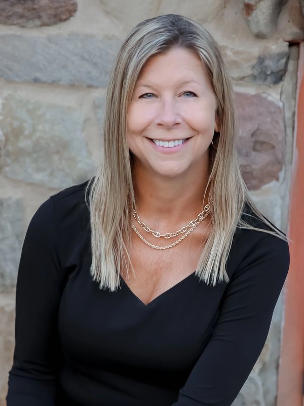 Smiling woman with blonde hair in a black top against a stone wall background.