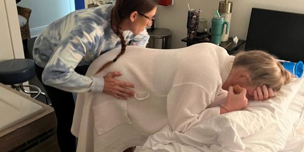 A woman supports another woman leaning on a bed, comforting her.