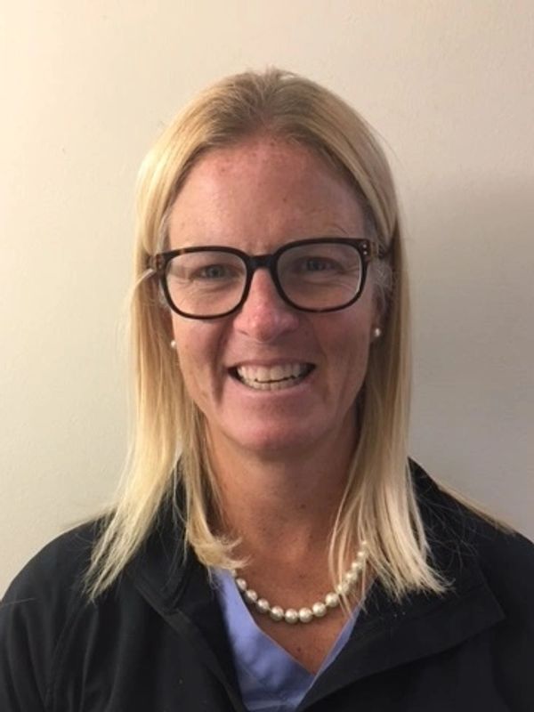 Smiling woman with glasses and pearl necklace against a plain wall.