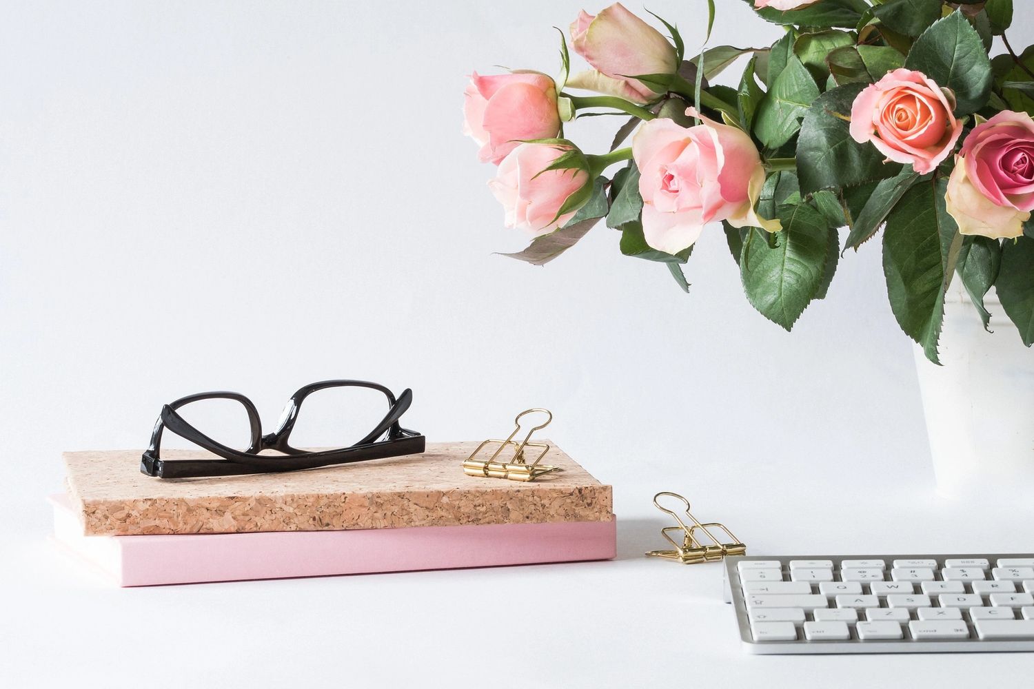 Glasses on a pile of books, next to pink roses in a vase and a keyboard. Bulldog clips.