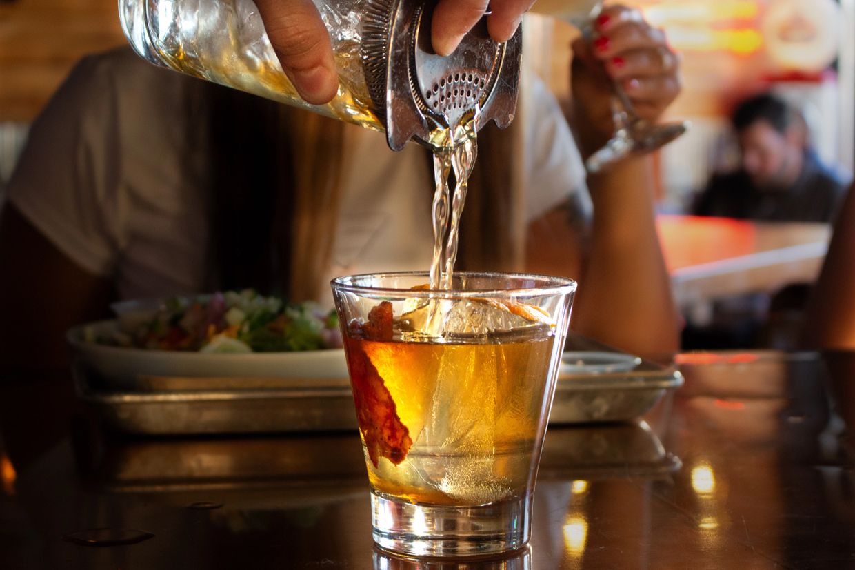 A cocktail is being poured into a glass with a large ice cube and garnish.