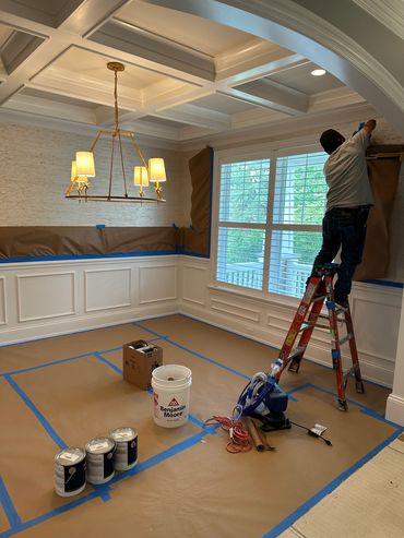 Room being prepared for painting with protective coverings and painter on ladder.