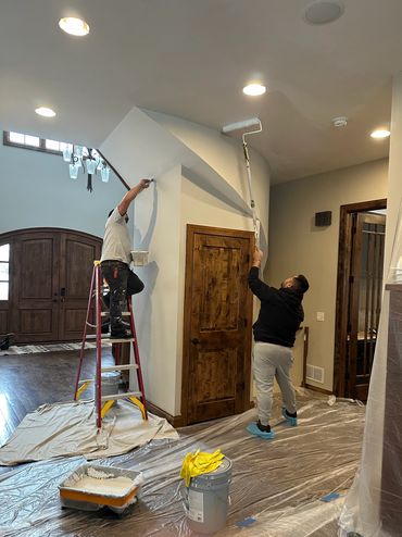 Two men painting a wall inside a house with ladders and rollers.