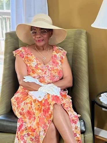 Elderly woman in floral dress and wide-brimmed hat sitting gracefully.