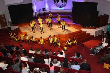 Children performing a lively dance in colorful costumes on stage before an audience.