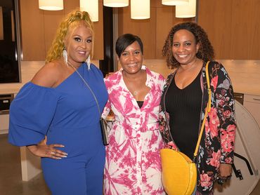 Three women smiling and posing indoors in colorful outfits.