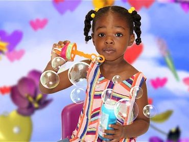 A young girl blowing bubbles with colorful flowers in the background.