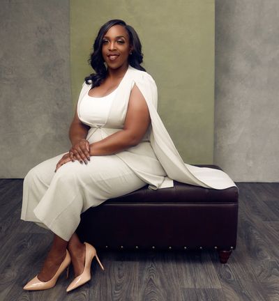 Confident woman in a white outfit sitting on a dark leather ottoman.