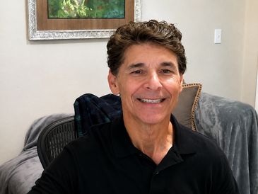 Smiling man in a black polo shirt sitting in a cozy room.