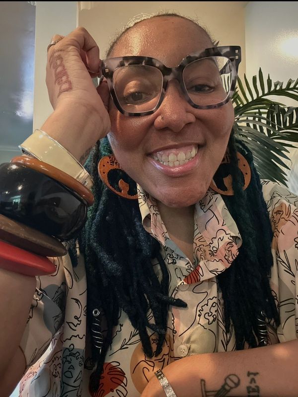 Smiling person with large glasses, colorful bracelets, and artistic shirt.