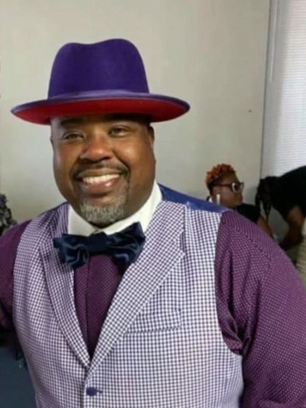 Man wearing a purple hat and checkered vest smiling.