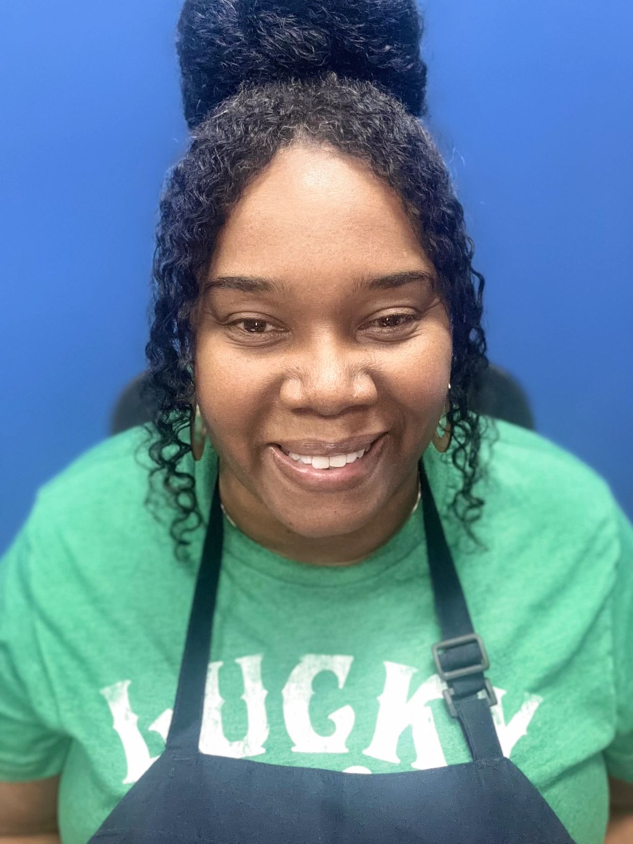 Smiling woman wearing a green shirt and black apron against a blue background.