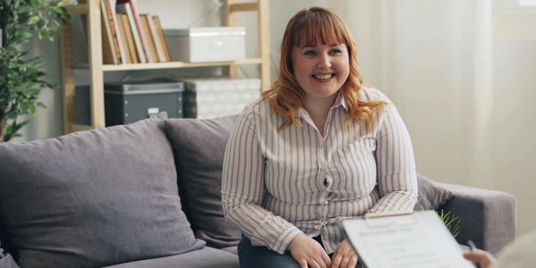 A smiling woman sits on a couch during a conversation.