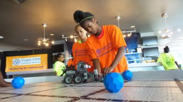 Two girls in orange shirts working on a robot at a STEM event.