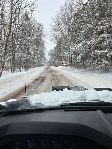 Snow-covered road viewed from inside a vehicle with snow on the hood.