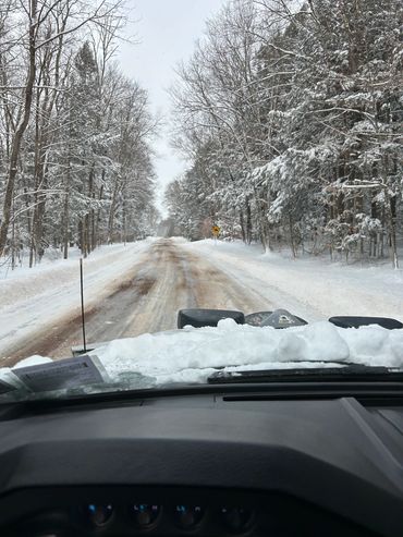 Snow-covered road viewed from inside a vehicle with snow on the hood.