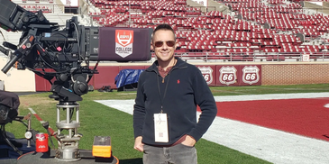 Man stands beside a large TV camera on a football field with empty stands.