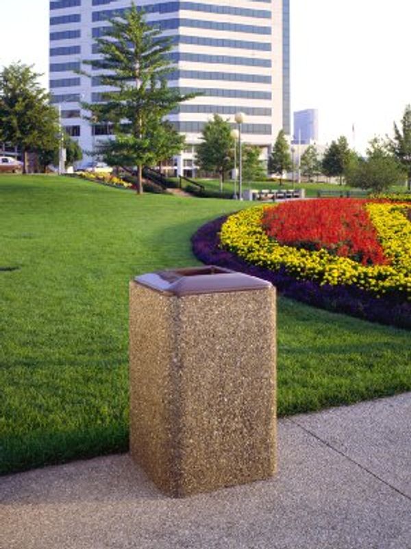 A trash bin in a city park with colorful flower beds and tall buildings.