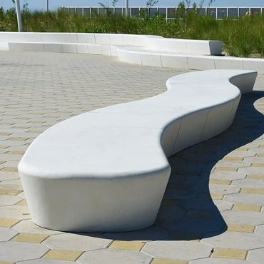 Curved white concrete bench on patterned pavement in a landscaped urban area.