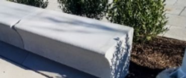 Concrete bench with a crack along the top, placed on pavement near bushes.