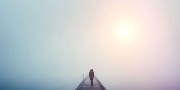 A person walks alone on a foggy pier towards a bright light.