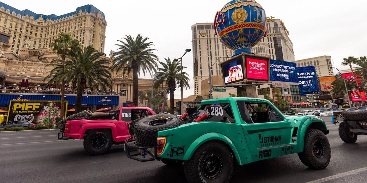 Colorful off-road trucks on a busy Las Vegas street with iconic landmarks in the background.