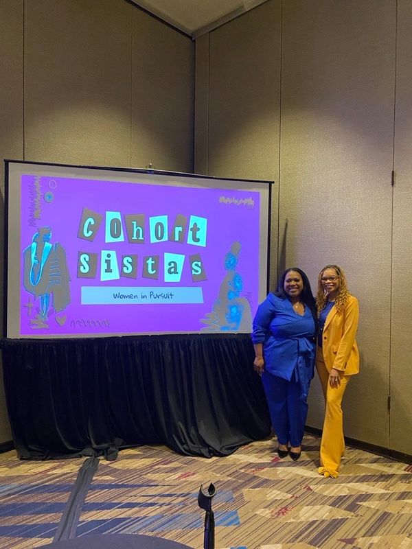 Two women stand beside a screen with "Cohort Sistas: Women in Pursuit" displayed.