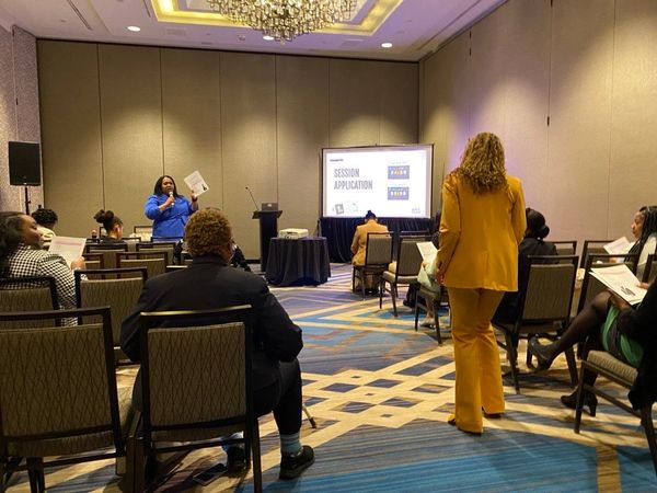 Woman in blue speaking at a session with attendees seated and one standing in yellow.