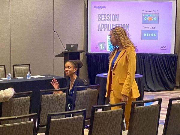 Two women interacting in a conference room with a presentation screen behind them.