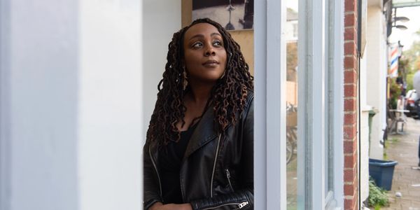 A woman with braided hair stands thoughtfully by a window in a leather jacket.