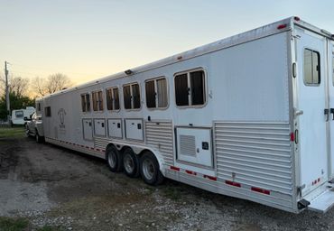 Large white horse trailer parked on gravel near trees at sunset.
