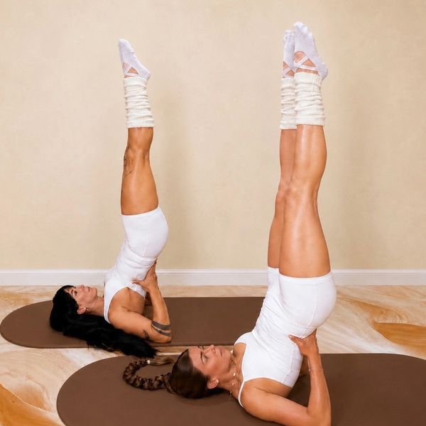 Two women practicing yoga shoulder stand pose indoors on mats.