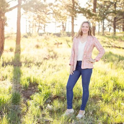 Woman smiling in a sunlit forest clearing wearing a pink blazer and jeans.