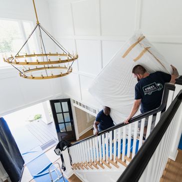 Two movers carry a large wrapped mattress down a staircase in a bright home.
