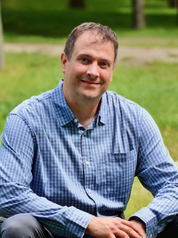Man in a blue checkered shirt smiling outdoors.