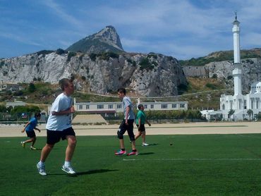 People playing cricket on a green field with a rocky mountain and mosque in the background.