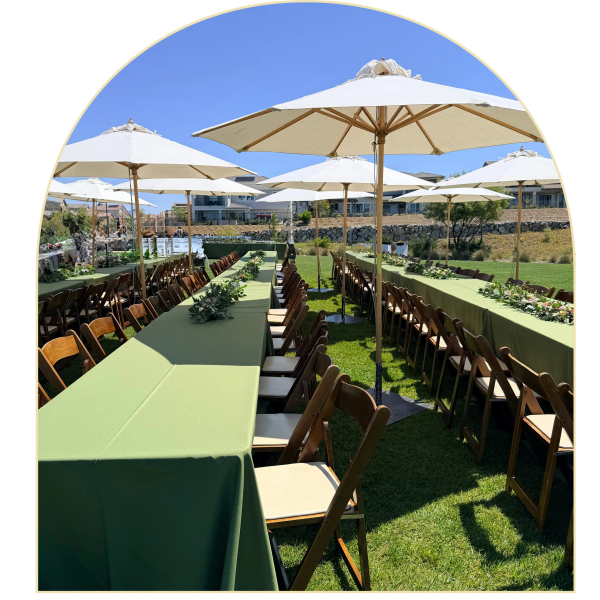 Outdoor event setup with green tablecloths, wooden chairs, and white umbrellas under a clear blue sky.